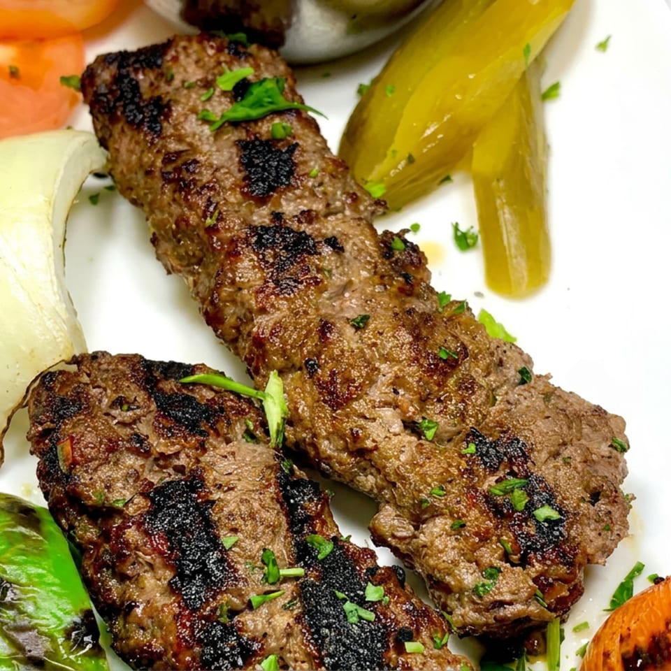 Grilled beef kebabs garnished with herbs, served with charred onion, green pepper, tomato, and pickled vegetables on a white plate.